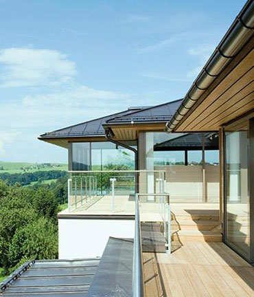 Een modern huis met een groot balkon en glazen balustrade, omgeven door een groen landschap onder een blauwe hemel.