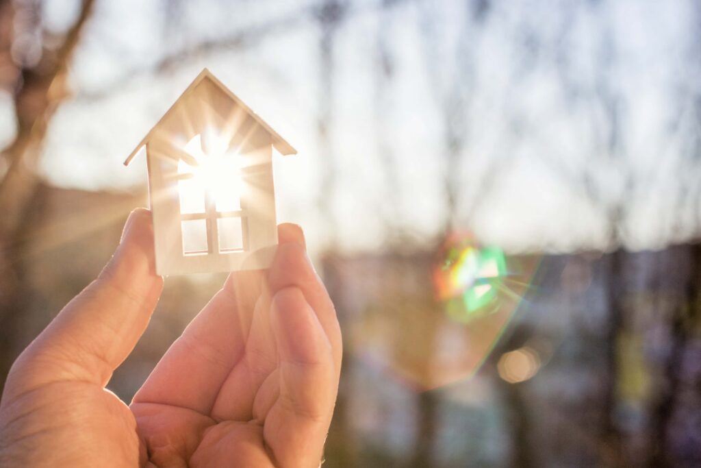 Een hand houdt een klein huismodel tegen het zonlicht met een wazige achtergrond van bomen.