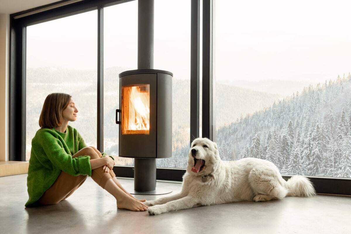 Een vrouw zit ontspannen voor een open haard met haar witte hond, terwijl ze door grote ramen uitkijkt op een besneeuwd landschap.