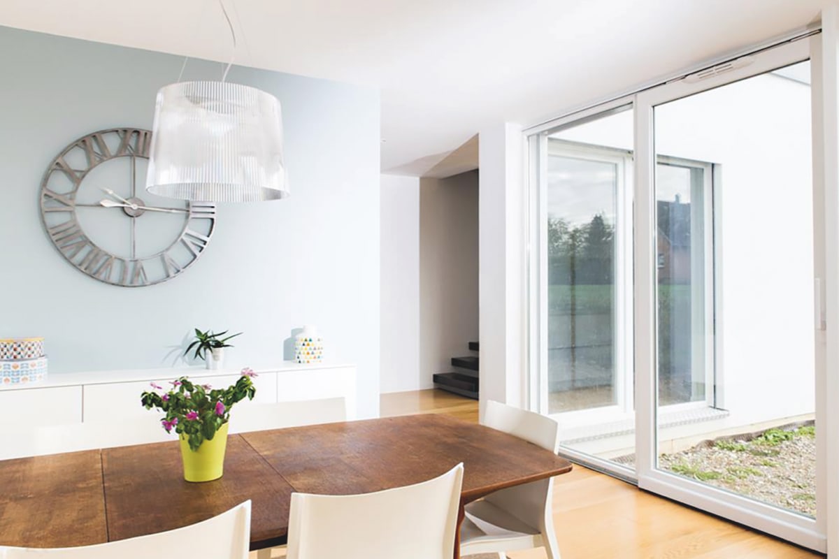 Een moderne eetkamer met een grote houten tafel, witte stoelen, een wandklok en een schuifdeur met uitzicht naar buiten.