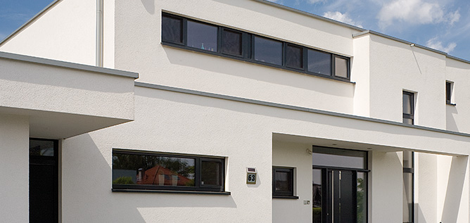 Modern wit huis met horizontale ramen, donkere kozijnen en een overhangend dak, onder een blauwe lucht met wolken.