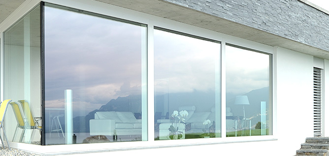 Modern huis met grote glazen schuifdeuren, witte kozijnen en uitzicht op bergen, met een lichte woonkamer en terrasmeubilair zichtbaar.