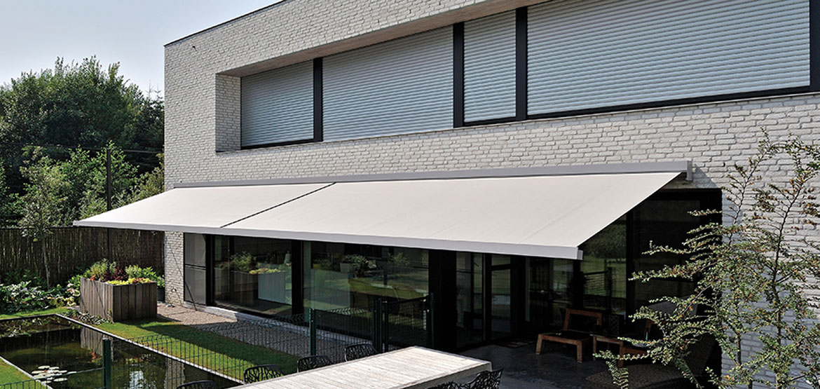 Wit bakstenen gebouw met gesloten rolluiken, glazen ramen en deuren, witte zonwering en een terras met een tuin.