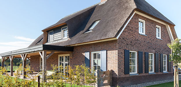 Traditioneel bakstenen huis met rieten dak, grote ramen, overdekt terras en groene tuin.