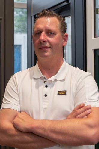 Man in wit poloshirt met gekruiste armen, staand in een moderne binnenruimte met een achtergrond van glas en zicht naar buiten.