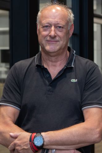 Man van middelbare leeftijd in donker poloshirt met horloge en armband, staand in een moderne glazen binnenruimte.