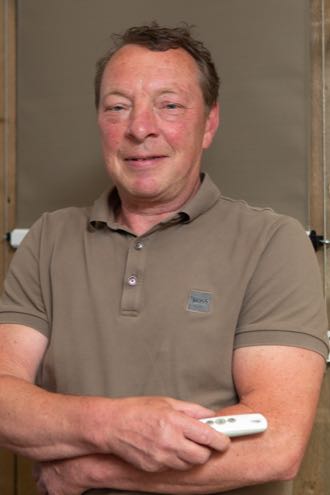 Oudere man in beige poloshirt met afstandsbediening in de hand, glimlachend in een neutrale binnenruimte.