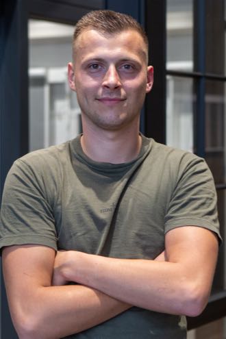 Jonge man met groen T-shirt en gekruiste armen, glimlachend voor een moderne glazen achtergrond.