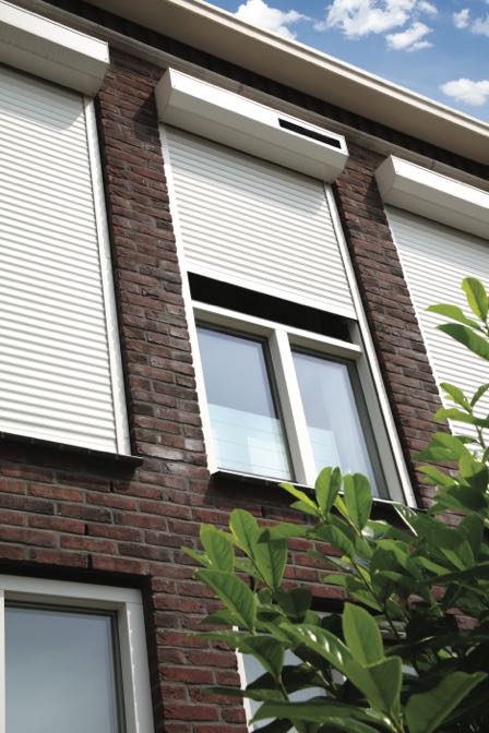 Bakstenen gevel van een gebouw met gesloten witte rolluiken en een geopend raam, omringd door groene planten.