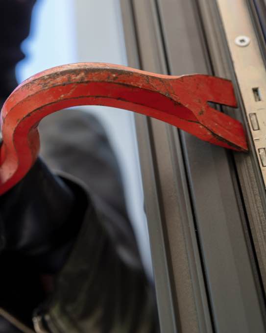 Close-up van een breekijzer in de hand van een persoon die een raam probeert open te breken, gekleed in zwarte kleding en handschoenen.