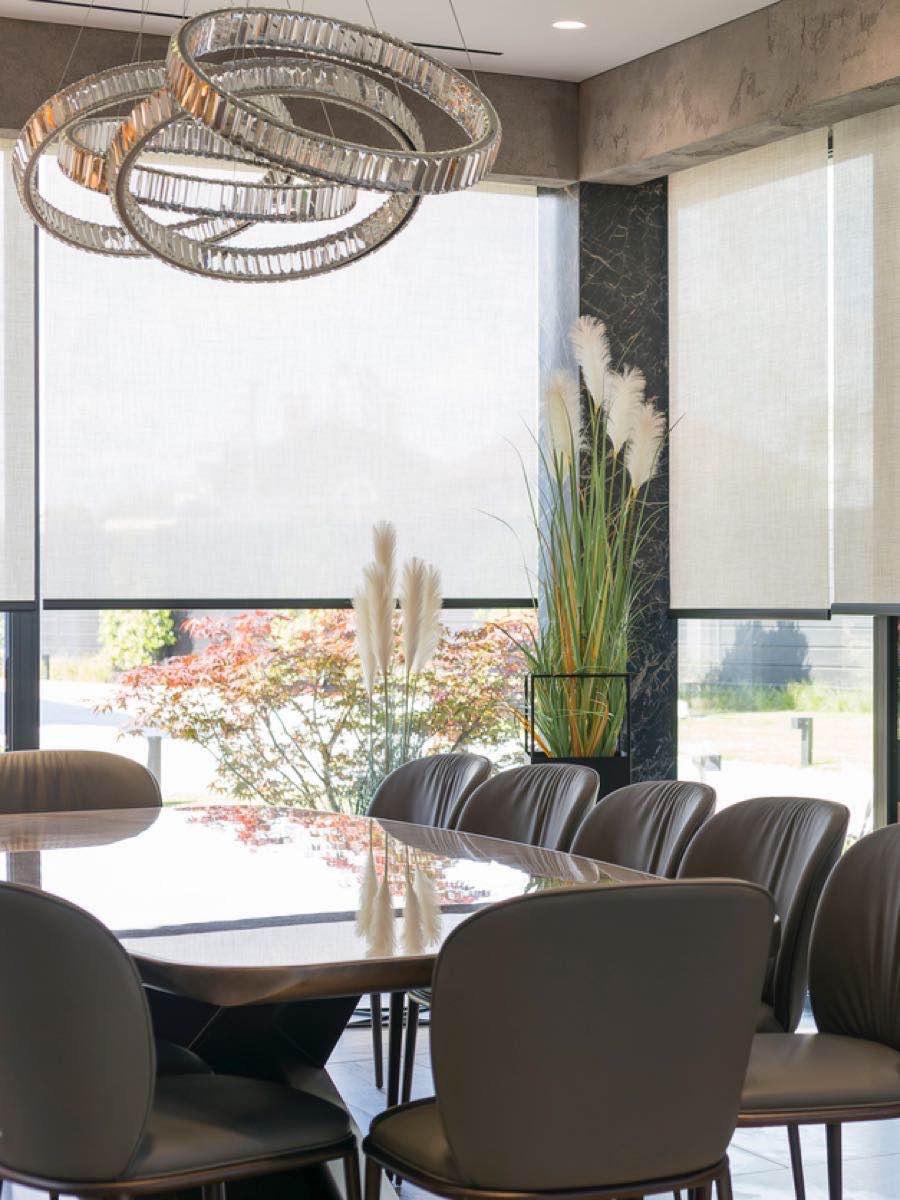 Moderne eetkamer met een elegante ronde tafel, leren stoelen, decoratieve planten en grote ramen met jaloezieën die zacht licht binnenlaten.