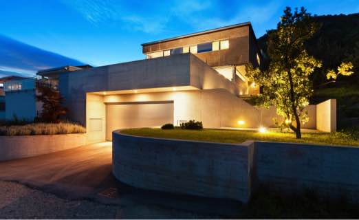 Modern huis met een betongevel en een verlichte oprit bij schemering, omgeven door natuur.