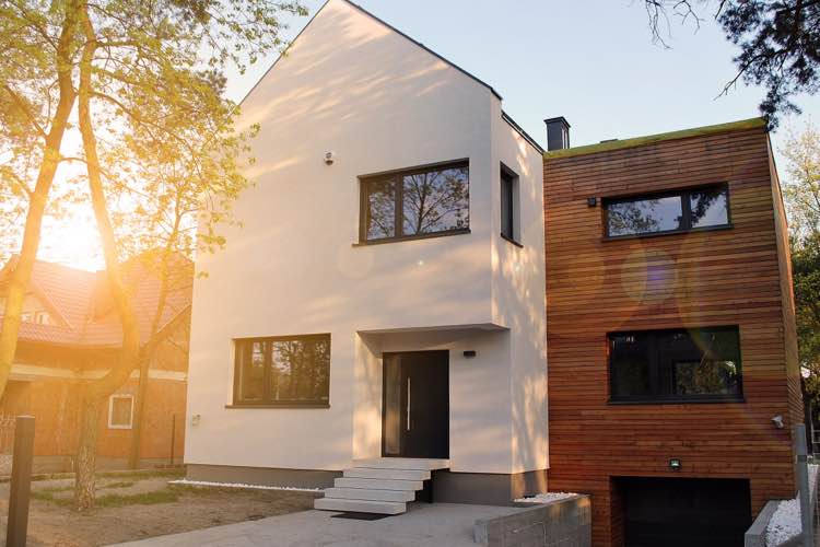 Een modern woonhuis met een lichte gevel en houtelementen, omgeven door bomen en zonlicht.