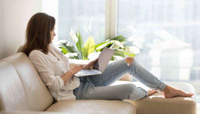 Jonge vrouw zit op een bank, werkt ontspannen met een laptop voor een groot raam