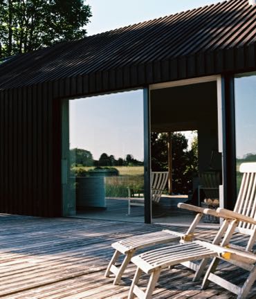 Modern huis met een donkere houten gevel en grote schuifdeuren, met daarvoor een terras met ligstoelen.